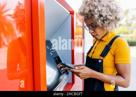 Femme utilisant un téléphone portable à un distributeur automatique de billets Banque D'Images