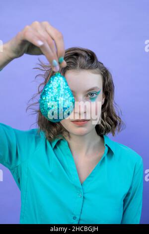 Fille portant une chemise en forme de turquoise tenant une poire pailletée devant un fond lavande Banque D'Images