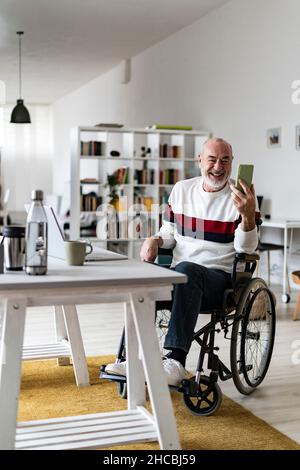 Homme d'affaires senior en fauteuil roulant souriant sur appel vidéo via smartphone au bureau à domicile Banque D'Images