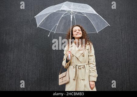 Photo horizontale d'une jeune femme attrayante portant un trench coat tenant un parapluie debout à l'extérieur contre un mur gris regardant la caméra Banque D'Images