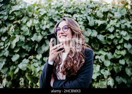 Femme d'affaires souriante envoyant la messagerie vocale via un smartphone devant la haie Banque D'Images