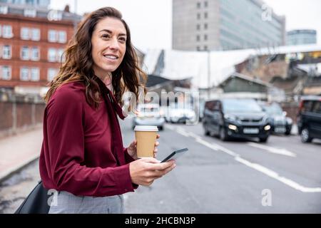 Femme d'affaires souriante attendant un taxi tenant un téléphone portable et une tasse de café dans la rue de la ville Banque D'Images