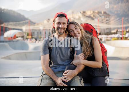 Couple souriant avec sacs à dos assis ensemble au parc de skateboard Banque D'Images