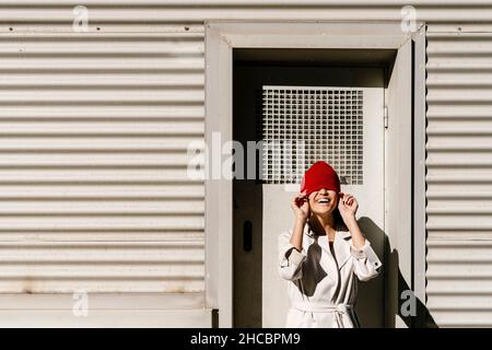 Jeune femme souriante couvrant le visage avec un chapeau en tricot par beau temps Banque D'Images