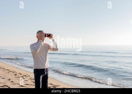 Homme regardant à travers des jumelles se tenant sur la plage Banque D'Images