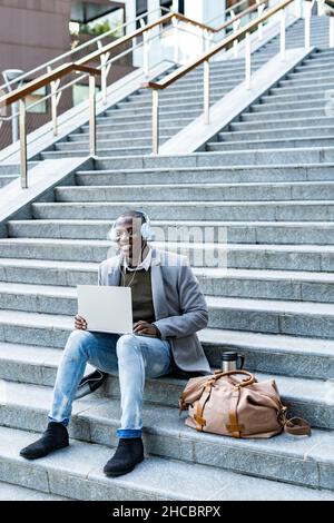 Homme d'affaires souriant avec un casque à l'aide d'un ordinateur portable placé sur un escalier Banque D'Images