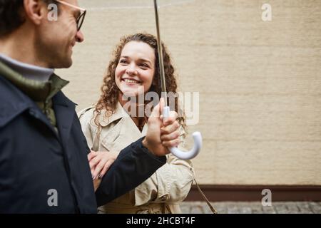 Photo de taille moyenne horizontale de couple moderne passant du temps ensemble en plein air marchant le jour de la pluie et souriant, homme tenant un parapluie Banque D'Images