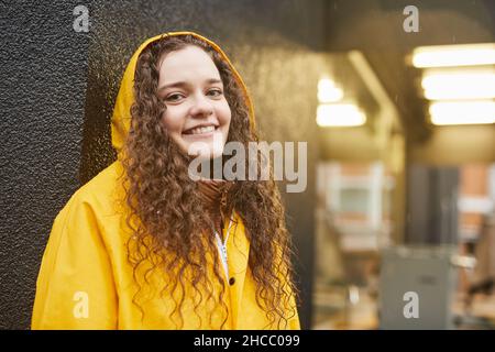 Gros plan horizontal moyen d'une femme caucasienne joyeuse portant un imperméable jaune debout sous la pluie souriant à l'appareil photo Banque D'Images