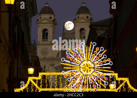 Italie, Rome, 18 décembre 2021 : la pleine lune apparaît parmi les lumières de Noël dans la rue Condotti, église de Santissima Trinita' dei Monti à backgrou Banque D'Images