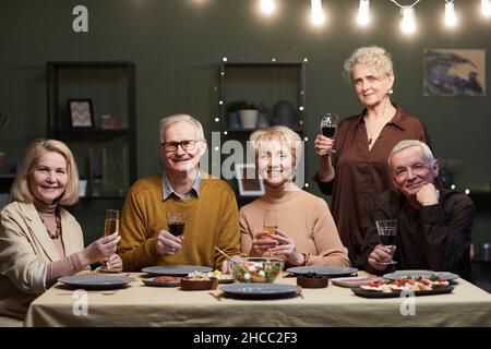 Un groupe de cinq amis seniors se sont réunis à la table à manger, tenant des verres de vin et de champagne tout en regardant l'appareil photo et en souriant Banque D'Images