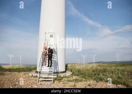 Partenaires multiraciaux debout sur les escaliers près de l'éolienne et regardant sur l'écran de la tablette numérique.Deux travailleurs de l'industrie qui se sont mis à faire fonctionner le système d'exploitation écologique à l'aide de gadgets modernes. Banque D'Images