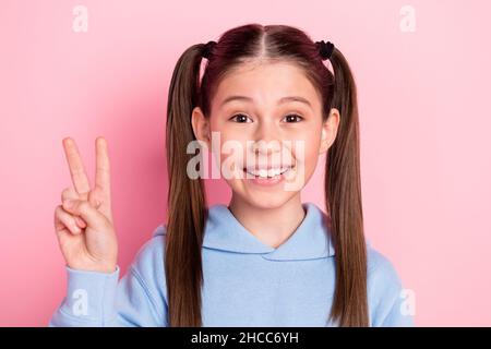 Photo portrait d'une écolière heureuse montrant un geste en V-sign souriant dans une tenue bleue isolée sur fond rose pastel Banque D'Images