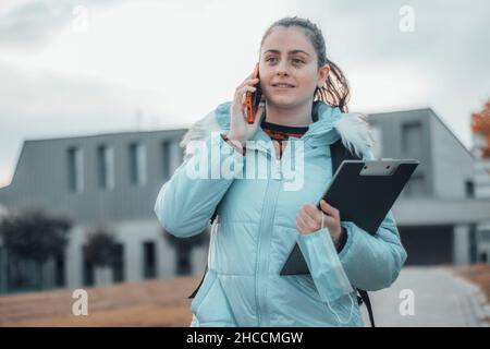 Jeune fille étudiante en médecine à l'extérieur se reposant à l'évidement tout en appelant sur le téléphone portable avec joie avec ses notes et dossier Banque D'Images