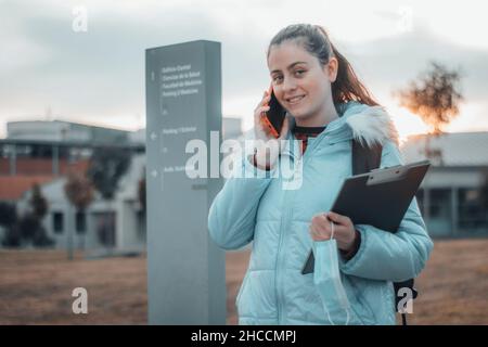 Une adolescente sombre qui fait un appel téléphonique à l'université de médecine avec un rire heureux comme elle tient ses notes et dossier Banque D'Images