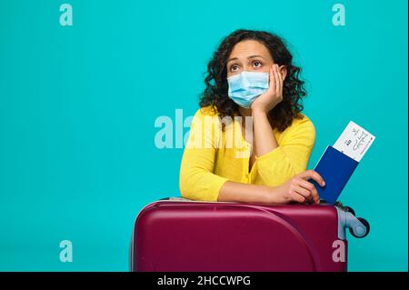 Charmante femme bouleversée en tissu jaune, portant un masque médical de protection, tient un passeport avec un billet et une carte d'embarquement, se penche sur ses bagages et regarde dr Banque D'Images