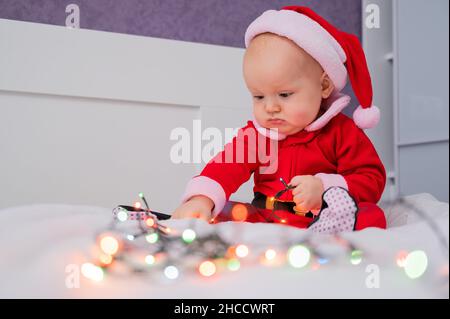 Bébé garçon portant au père noël assis sur le lit jouant avec des lumières. Banque D'Images