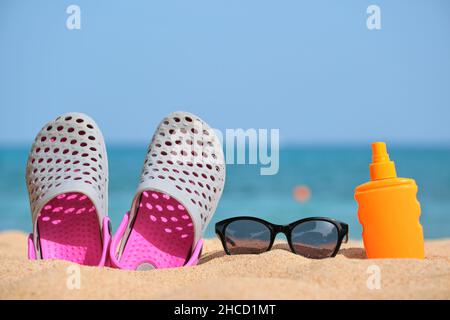 Gros plan sur des chaussures de sabots, de la crème solaire et des lunettes de soleil noires de protection sur une plage de sable au bord de la mer tropicale par temps chaud et ensoleillé. Concept de vacances d'été. Banque D'Images