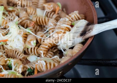 Gros plan de pâtes de blé entier, de légumes et de fromage fondu cuisant dans une casserole en céramique orange sur une cuisinière et étant remué par une cuillère de cuisson en bois Banque D'Images