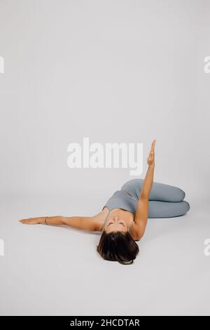 Brunette est engagée dans le sport.La jeune femme fait de l'exercice de yoga ou de fitness, elle se repose sur le dos, soulève un bras, jambes pliées aux genoux, gris Banque D'Images