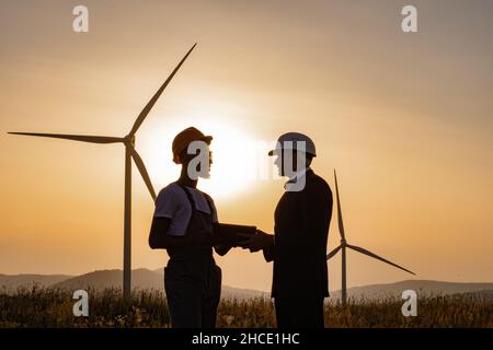 Silhouette d'un ingénieur afro-américain et d'un inspecteur indien utilisant une tablette numérique lors d'une réunion à l'extérieur.Deux partenaires debout sur le terrain avec des éoliennes pendant un incroyable coucher de soleil d'été. Banque D'Images