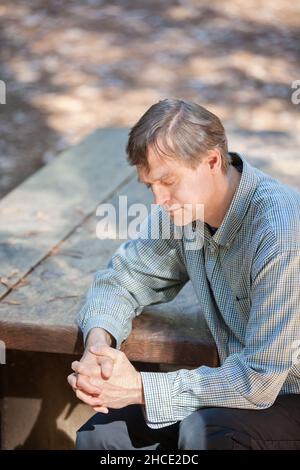 Homme caucasien dans les années cinquante assis seul à table en bois priant dans la forêt Banque D'Images