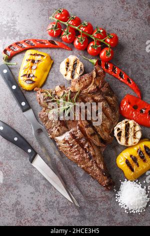 Gros steak juteux en T cuit sur le gril servi avec des légumes grillés sur fond gris en béton.Vue verticale du dessus Banque D'Images