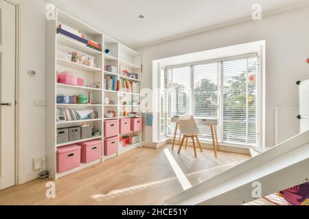 placard blanc avec divers livres et fournitures sur les étagères avec des conteneurs colorés dans la salle d'étude lumineuse avec table près de la grande fenêtre Banque D'Images