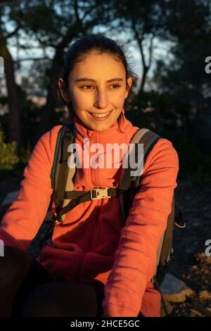 Randonneur féminin souriant avec sac à dos à la vue sur la distance tout en étant assis dans la nature avec la lumière du soleil pendant le trekking Banque D'Images