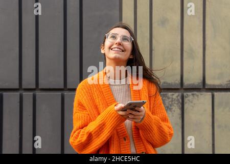 Jeune femme gaie en lunettes et gilet orange chaud défilant médias sociaux sur smartphone debout sur un mur de béton Banque D'Images