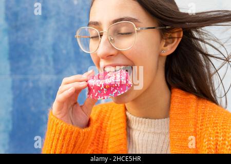 Jeune femme positive en lunettes et gilet chaud tricoté mangeant savoureux donut rose arrosé avec les yeux fermés Banque D'Images