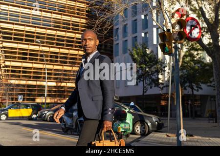 Homme d'affaires afro-américain sans émotion en vêtements formels avec porte-documents traversant une route asphaltée sur une rue ensoleillée avec des bâtiments dans la ville Banque D'Images