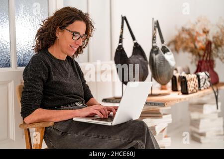 Contenu femme designer parcourant un netbook moderne tout en étant assis près d'une table avec divers sacs à main tendance dans la salle d'exposition de conception légère Banque D'Images