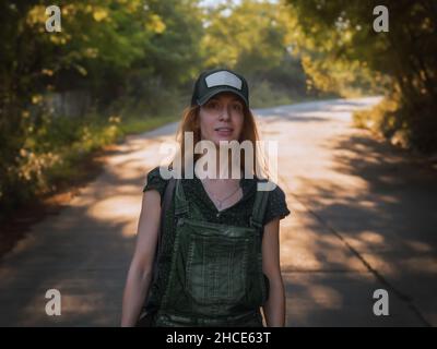 Une femme au Cap marche le long d'une route ombragée avec une forêt des deux côtés, le jour de l'été.Il y a un endroit pour une mocap sur la casquette Banque D'Images