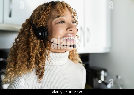 Une femme afro-américaine charmante et positive avec un casque à cheveux bouclés tout en se tenant dans une cuisine légère et en regardant loin Banque D'Images