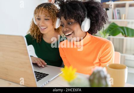 Des amies multiraciales positives écoutent et regardent des films sur un ordinateur portable, assis dans un casque dans la chambre pendant la journée Banque D'Images