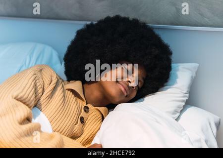 Jeune femme afro-américaine avec une coiffure afro et des yeux fermés dormant paisiblement sur un confortable lit blanc à la maison Banque D'Images