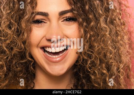 Portrait en gros plan de la jeune femme avec des cheveux bouclés souriant et regardant l'appareil photo tout en se tenant debout sur fond rose Banque D'Images