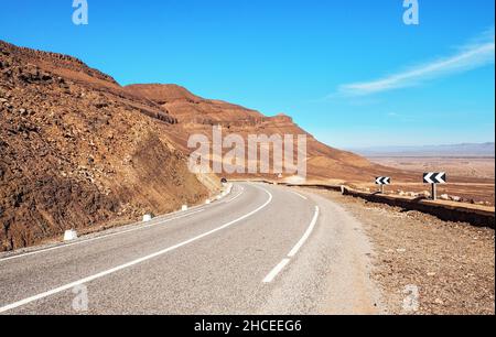 Courbe sur route asphaltée, petites montagnes de l'Atlas et buissons bas des deux côtés, ciel clair au-dessus - paysage typique dans le sud du Maroc Banque D'Images