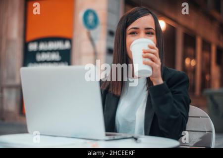 Une jeune femme souriante qui boit un café dans un café de la ville tout en travaillant avec son ordinateur portable Banque D'Images