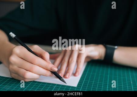 Gros plan des mains d'une femme designer méconnue qui marque avec un crayon sur du papier blanc couché sur un tapis de découpe en caoutchouc au bureau, mise au point sélective. Banque D'Images