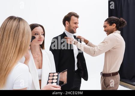 styliste et maquilleur afro-américains joyeux préparant de jeunes modèles élégants en studio photo Banque D'Images