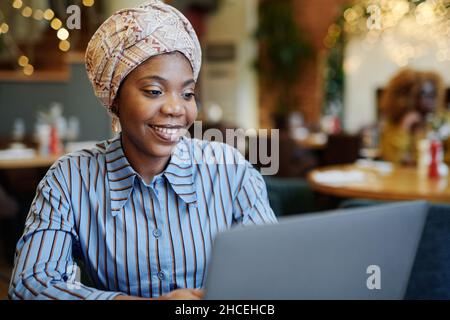 Jeune femme africaine assise à la table et souriante tout en travaillant sur un ordinateur portable dans un café Banque D'Images