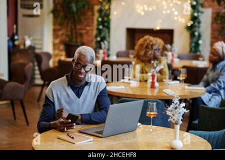 Un jeune homme d'affaires heureux lisant un message sur un téléphone portable tout en étant assis à la table avec un ordinateur portable dans un café Banque D'Images