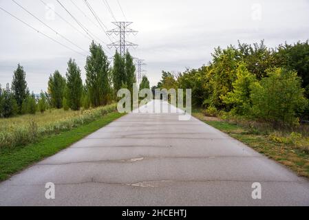 Sentier pavé déserté pour les pédalins et les cyclistes le long de la rive d'un lac. Des câbles haute tension aériens soutenus par de grands pylônes alignent le chemin. Banque D'Images