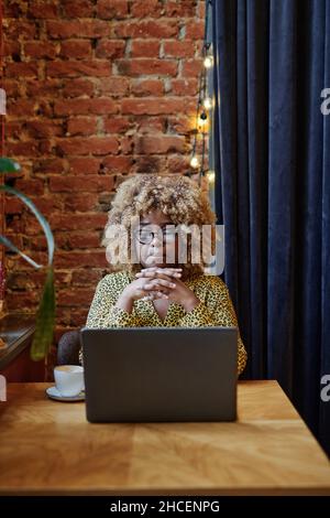 Femme d'affaires africaine aux cheveux bouclés assise à la table devant l'ordinateur portable dans un café et pensant au travail en ligne Banque D'Images