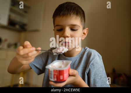 Un adorable garçon avec l'appétit mange du yogur avec une cuillère, des grimaces. Banque D'Images