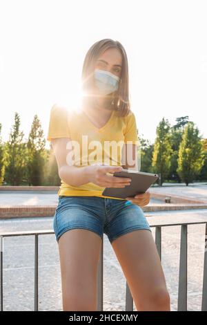 Jeune fille blonde de cheveux portant un t-shirt jaune, masque chirurgical, regardant son comprimé.Technologie, style de vie, détente, quarantaine, covid-19 et omicron var Banque D'Images