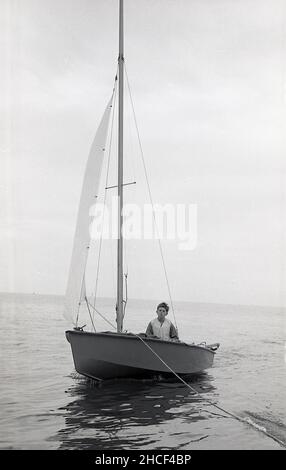 1960s, historique, un adolescent, portant un gilet de sauvetage de l'époque, à la conduite d'un petit voilier ou d'un canot pneumatique saling sur une zone calme de la mer, Angleterre, Royaume-Uni. Banque D'Images