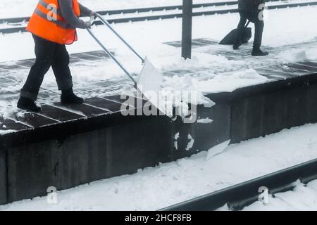 Une femme travaillant dans un gilet orange réfléchissant nettoie la neige avec une pelle spéciale au chemin de fer. Banque D'Images