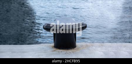 Un bollard noir sur la jetée du port, un fond d'eau de mer ondulé, un espace de copie.Poteau en métal lourd ancré sur un sol en béton de quai Banque D'Images